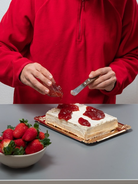 woman in red sweaters pours raspberry pudding into a white cake and next to it is a bowl full of strawberries.