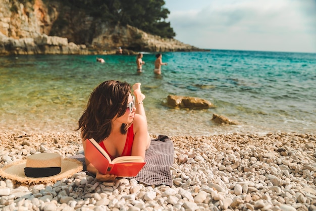 Woman in red swimsuit laying on blanket at sea beach reading book sun tanning