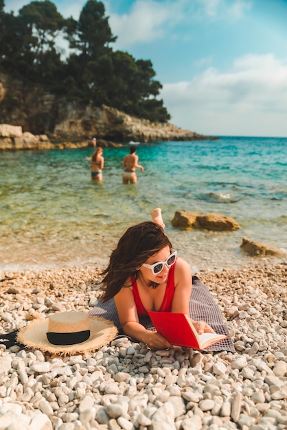 Woman in red swimsuit laying on blanket at sea beach reading book