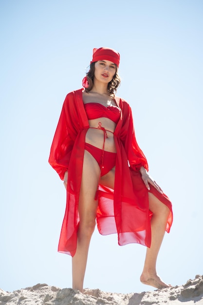 A woman in a red swimsuit and a red hat stands on a blue sky background.
