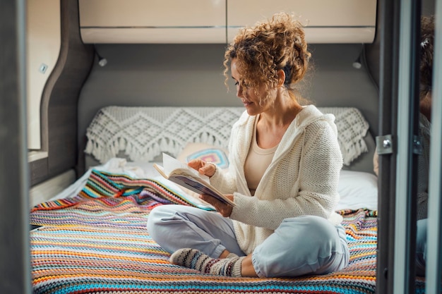 Woman relax sitting on the bed inside comfortable camper van\
reading a book indoor leisure activity and off grid van life travel\
people and holiday vacation on rented motor home serene female