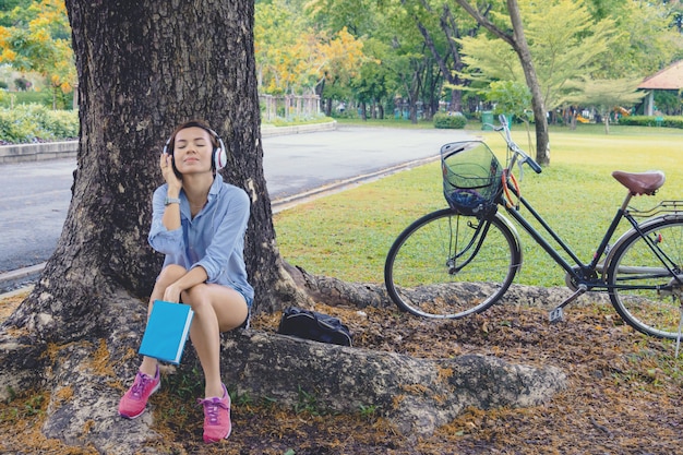 女性は公園で時間をリラックスします。彼女は大きな木の下で音楽を聴いて本を読んだ。