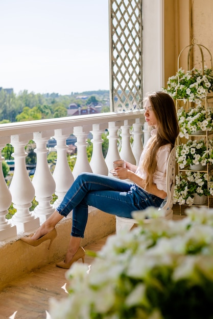 Foto donna che si distende e beve il caffè in terrazza, giornata di sole. giovane donna seduta su una sedia e rilassarsi a casa sul balcone, vicino ai fiori.