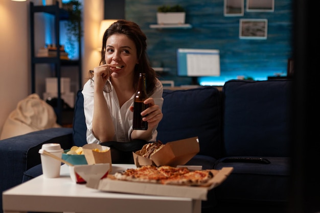 Woman relaxing on sofa watching television while drinking craft\
beer and eating potato chips in home living room. office worker\
enjoying a cold beer and tasty home deliverd fast food.