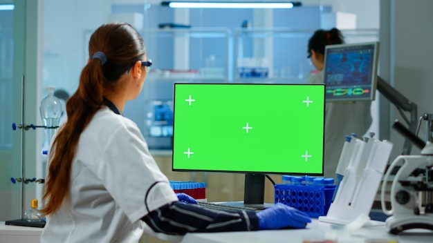 Woman researcher typing at computer with green screen mockup display, template in applied science laboratory. Engineer conducting experiments in background, examining vaccine evolution using high tech