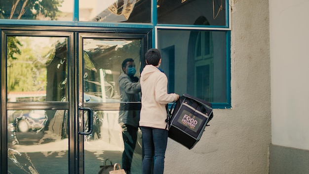 Woman restaurant courier letting fastfood order at front door, client picking up paperbag with meal lunch. Takeaway employee delivering food package during covid 19 pandemic, wearing facemask.