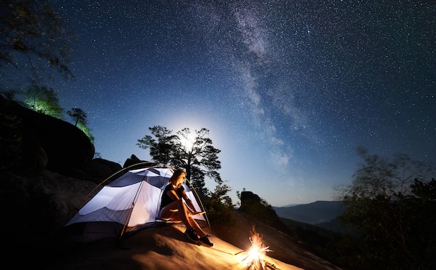 Woman resting beside camp, bonfire and tourist tent at night