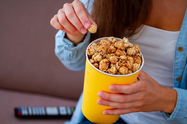 Женщина отдыхая на кресле и держа хрустящую коробку попкорна карамельки для закуски во время смотреть ТВ дома. Попкорн фильм