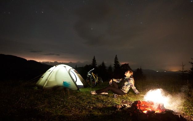 Woman resting at night camping near campfire, tourist tent, bicycle under evening sky full of stars