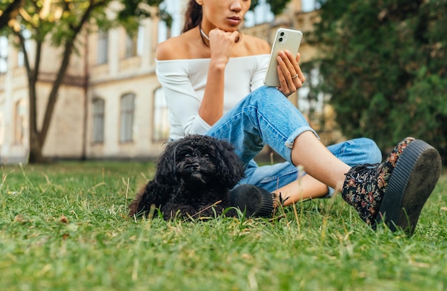 かわいい黒犬と一緒に公園の芝生に座って休んでいる女性は、スマートフォンのペットを使用しています