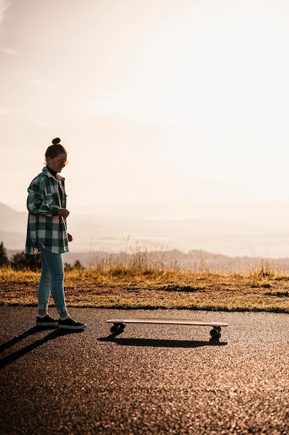 Foto la donna corre su strada dritta sul longboard al tramonto skater in abbigliamento casual si allena a bordo durante il tramonto serale con luce arancione ragazza tiene il longboard nelle mani