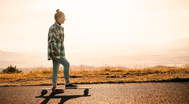 Woman rides at straight road on longboard at sunset time skater\
in casual wear training on board during evening sunset with orange\
light girl hold longboard in hands