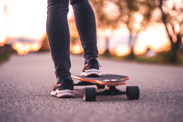女性は日没時にロングボードの直線道路に乗ります オレンジ色の光で夕方の日没時にボード上でカジュアルウェアのトレーニングをするスケーター 女の子は手でロングボードを保持します
