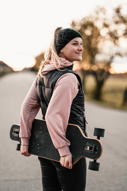 Woman rides at straight road on longboard at sunset time Skater in casual wear training on board during evening sunset with orange light Girl hold longboard in hands