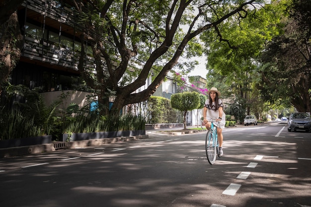 Woman riding a bicycle in the street with trees by the sides wearing white dress and hat