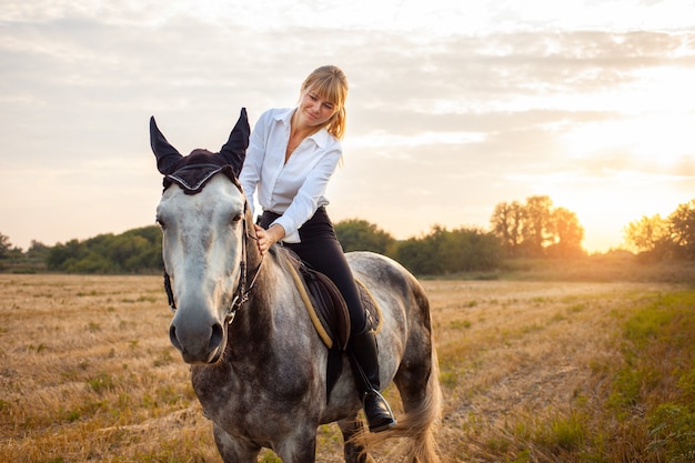 A woman riding a gray horse in a field at sunset. walking, horseback riding, rental. Beautiful background, nature outdoor. equestrian sport training. Copy space. love for the animal, friendship