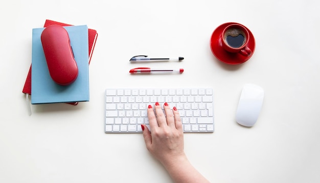 Woman right hand on wireless keyboard with wireless mouse, cup of coffee, pens, notebooks, glasses case on white table