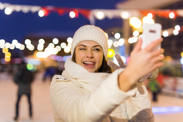 A woman on the rink is skating and taking selfie on smartphone. New Years Eve and Christmas. Fairy lights. Ice and snow mood concept. Winter sport.