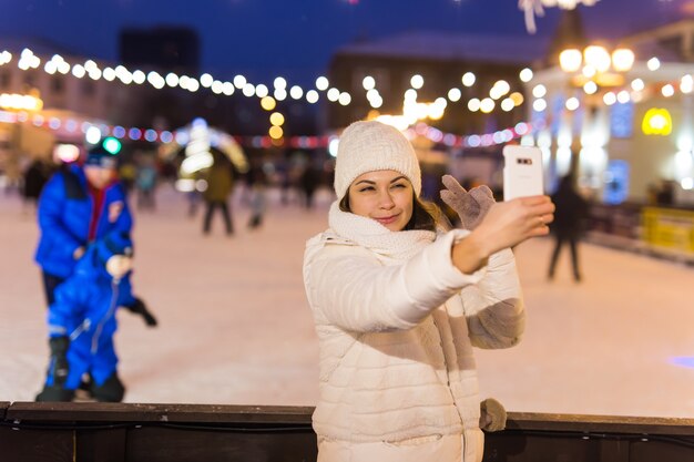 A woman on the rink is skating and taking selfie on smartphone. New Years Eve and Christmas. Fairy lights. Ice and snow mood concept. Winter sport.