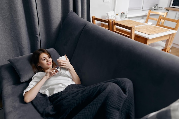 The woman in the room lies on the couch with a phone in her hand and a window in the background