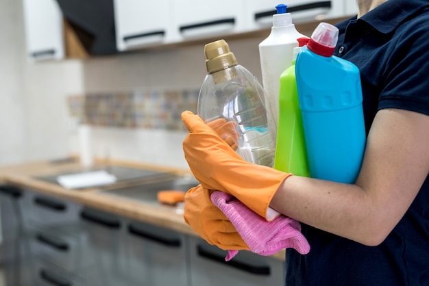 Woman in rubber gloves holding plastic bottles of washing liquid on the kitchen. Home cleaning concept.