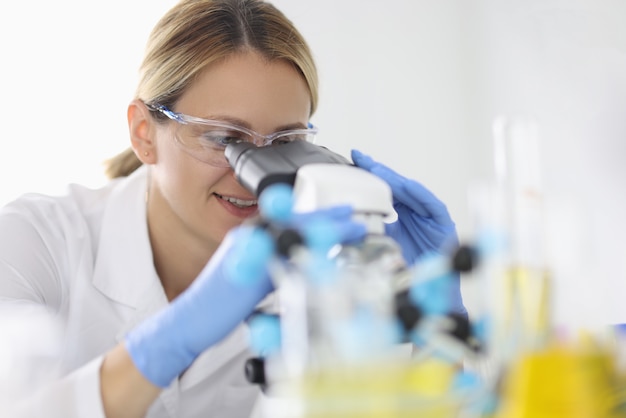 ゴム手袋と保護化学ガラスの女性は、実験室の肖像画の顕微鏡を通して見ています。臨床診断分析の概念を実施します。