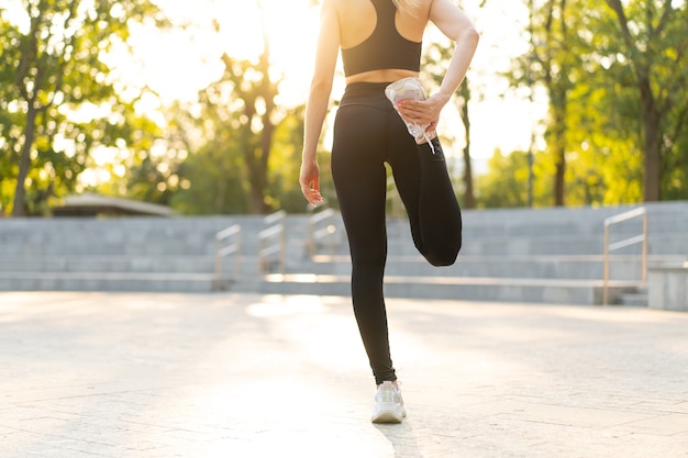 Woman runner stretching legs before exercising summer park morning Middle age athletic female warming up body before running Caucasian person warm up jogging Dressed white shirt shorts running track