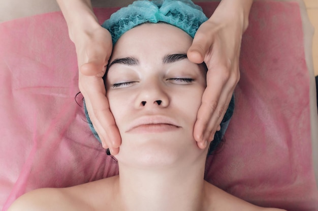 Woman's face in cosmetic cap and hands of beautician Cosmetic procedures washing facial massage