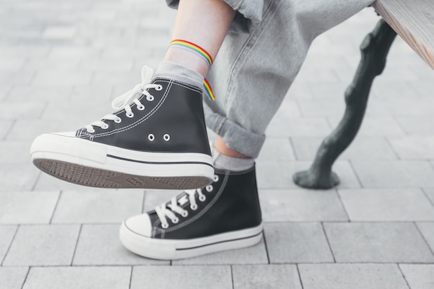 Foto piedi di donna in scarpe da ginnastica bianche e nere con braccialetto gay pride alla caviglia seduto su una panchina con colori insaturi