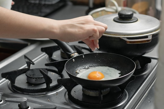 Photo a woman's hand adding salt to the egg she is frying in a frying pan on the burner of a gas stove