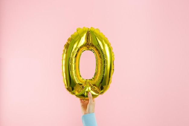 Woman's hand in blue jumper holding golden globe in the shape of number 0 on pink background. Copy space.