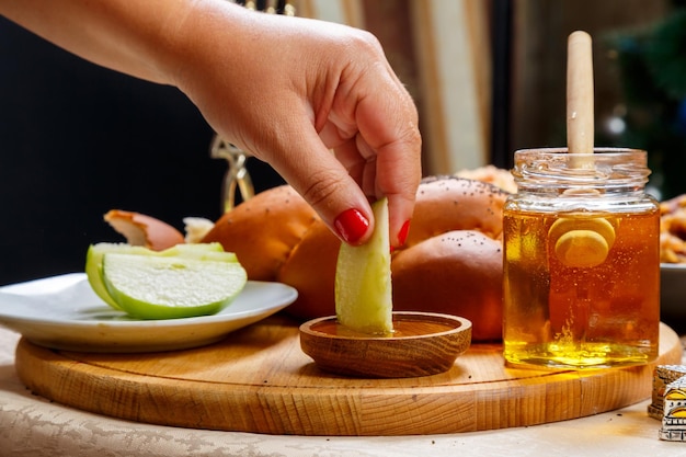 A woman's hand dips a piece of apple in honey in honor of the celebration of Rosh Hashanah near honey and challah and traditional food