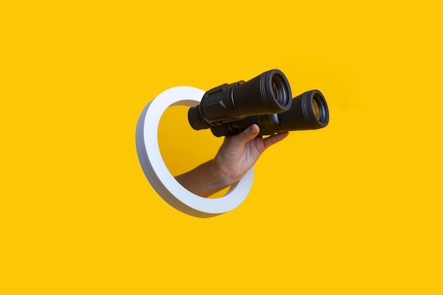 Photo woman's hand holding binoculars in a hole on a yellow background.