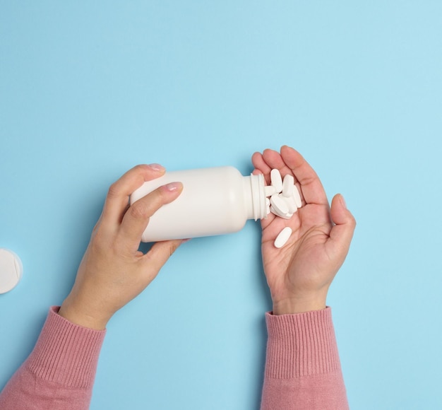 A woman's hand holding a plastic bottle and white oval pills on a blue background top view