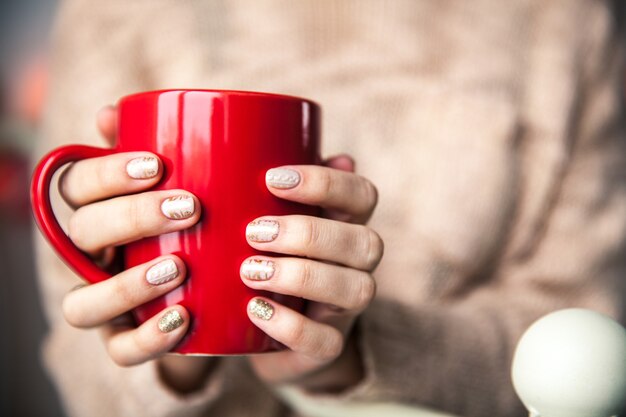 コーヒーの赤いカップを持っている女性の手。美しい冬のマニキュア付き。