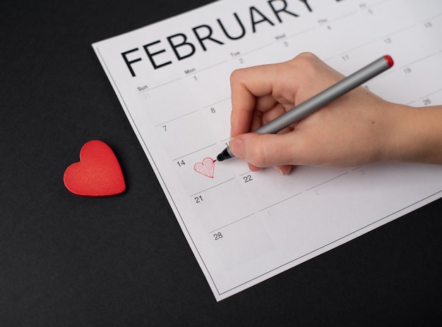 Woman's hand holding a red pen above the February 14 on the paper calendar and red wooden heart on the dark background