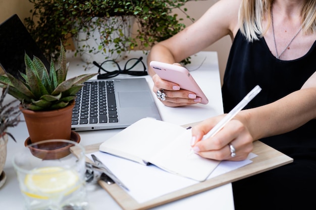 Woman's hand holding smartphone and making notes in notepad in cozy summer office with laptop