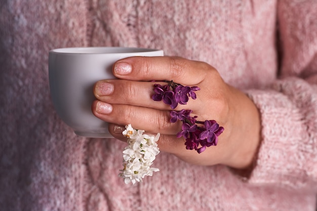 Woman's hand holding a white cup of coffee. With a beautiful lilac flowers. Drink, fashion, morning