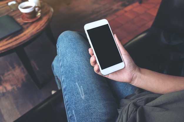 woman's hand holding white mobile phone with blank black desktop screen on thigh while sitting in cafe