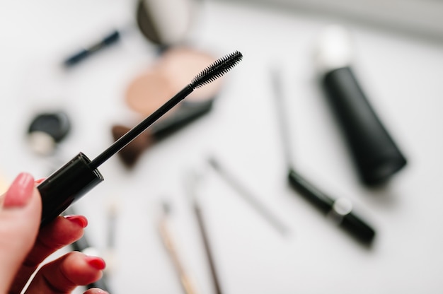 The woman's hand holds a brush for mascara on a blurry surface of different cosmetics