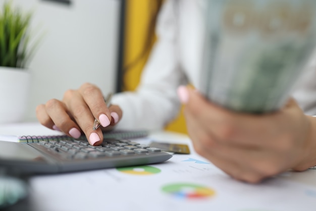 Woman's hand holds dollars and pen with the other hand and types numbers on calculator.