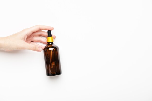 a woman's hand holds a glass brown bottle with pipette with essential oil on white background. aromatic cosmetic product for skin and hair care. moisturizing beauty product with organic natural oils.