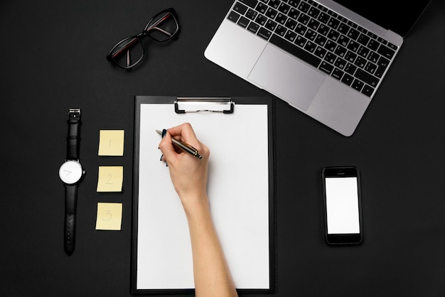 A woman's hand holds a pen and writes on a clean sheet of paper. Black office desk with laptop, phone and supplies. Yellow papers for notes with one, two, three numbers.