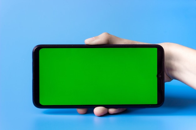 Photo the woman's hand holds the smartphone in a horizontal position with a green screen on a blue background. chroma key. mock up