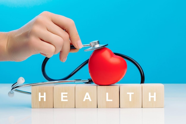 A woman's hand holds a stethoscope and touches a red heart that lies on top of wooden cubes with the inscription HEALTH