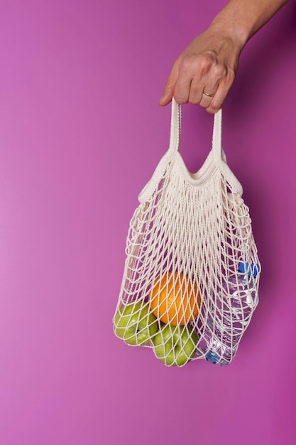 A woman's hand holds a string bag with oranges, apples and a bottle of water