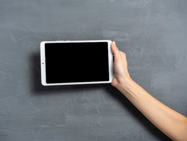 A woman\'s hand holds a white tablet on a gray textured\
background. place to insert on the screen of an electronic\
tablet.