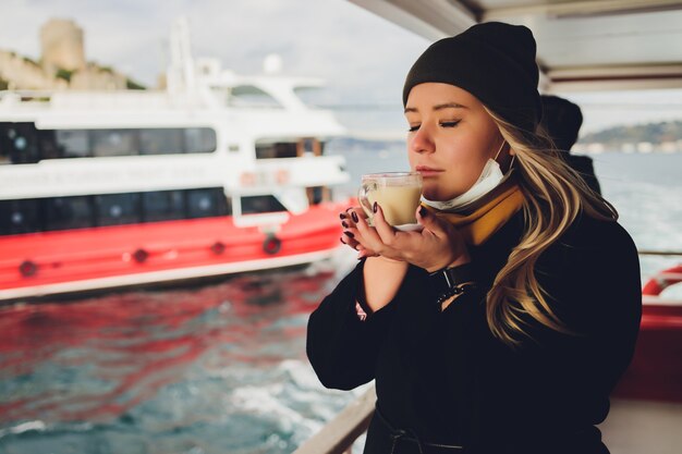 La mano di una donna tiene in mano una tazza bianca di bevanda calda a base di latte con cannella chiamata turca salep sahlep sullo sfondo dell'acqua increspata e della nebbiosa torre della fanciulla in lontananza, istanbul.