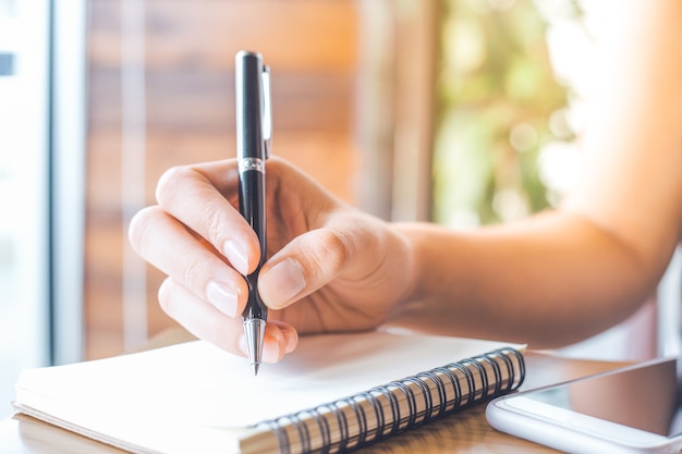 Woman's hand is writing on a blank notepad with a pen on a wooden desk