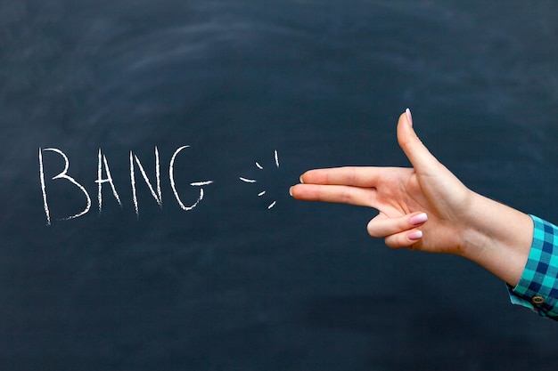 A woman's hand makes a shooting gesture with her hand, the inscription BANG on a chalkboard